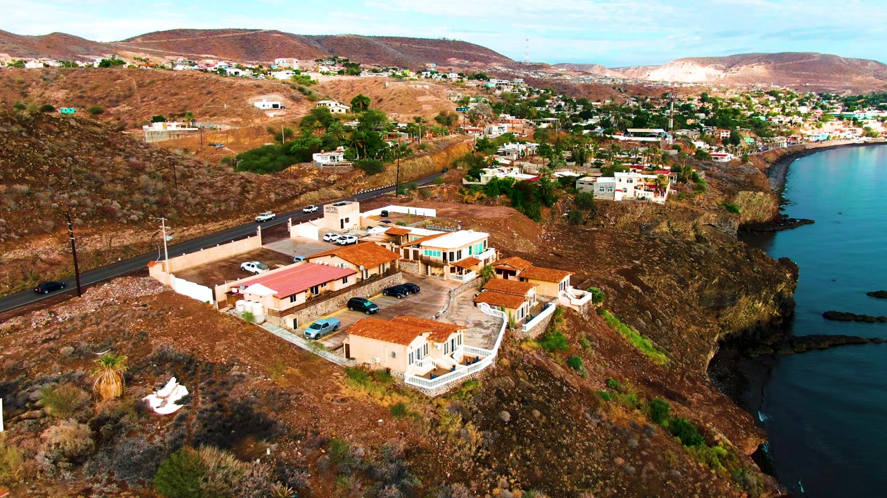 Aerial frame in Santa Rosalia showing the camera closer to the red-tiled building cluster, with parked cars in the lot, a road behind, ocean at right, and no visible people.