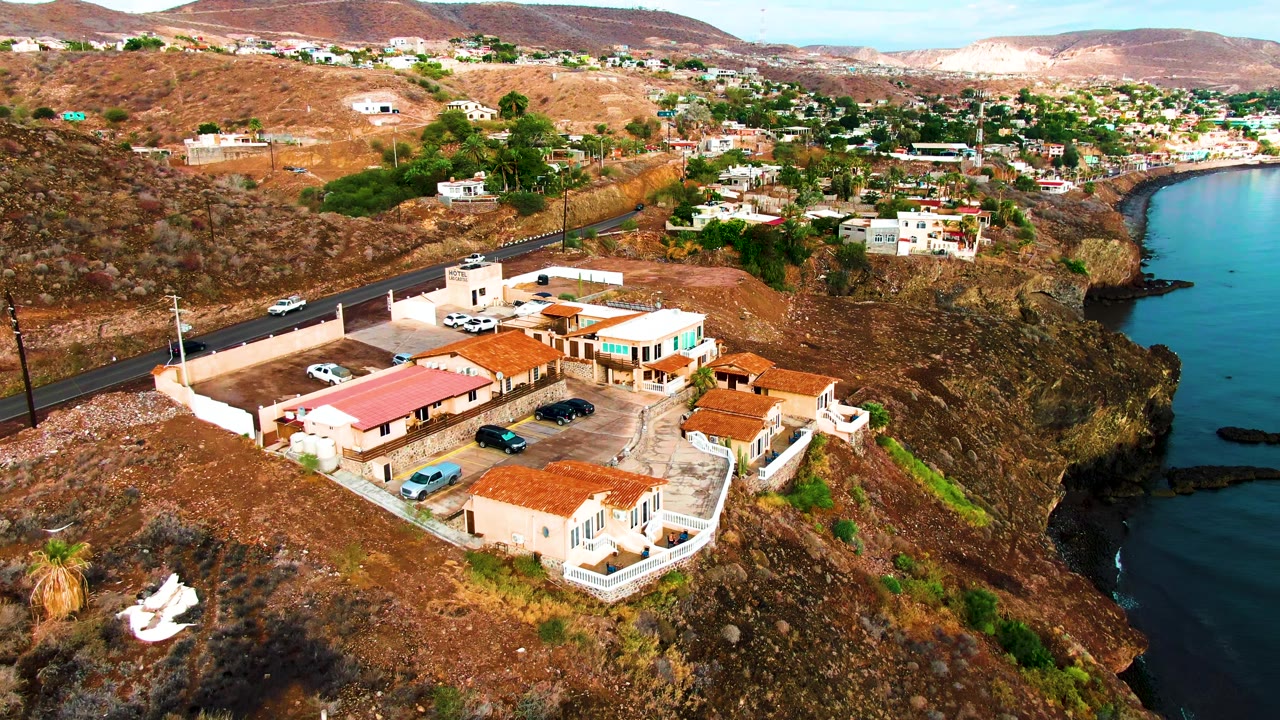 Santa Rosalia aerial frame with the building compound centered lower in the image, dry slopes rising behind it, a curved shoreline to the right, and no visible people.