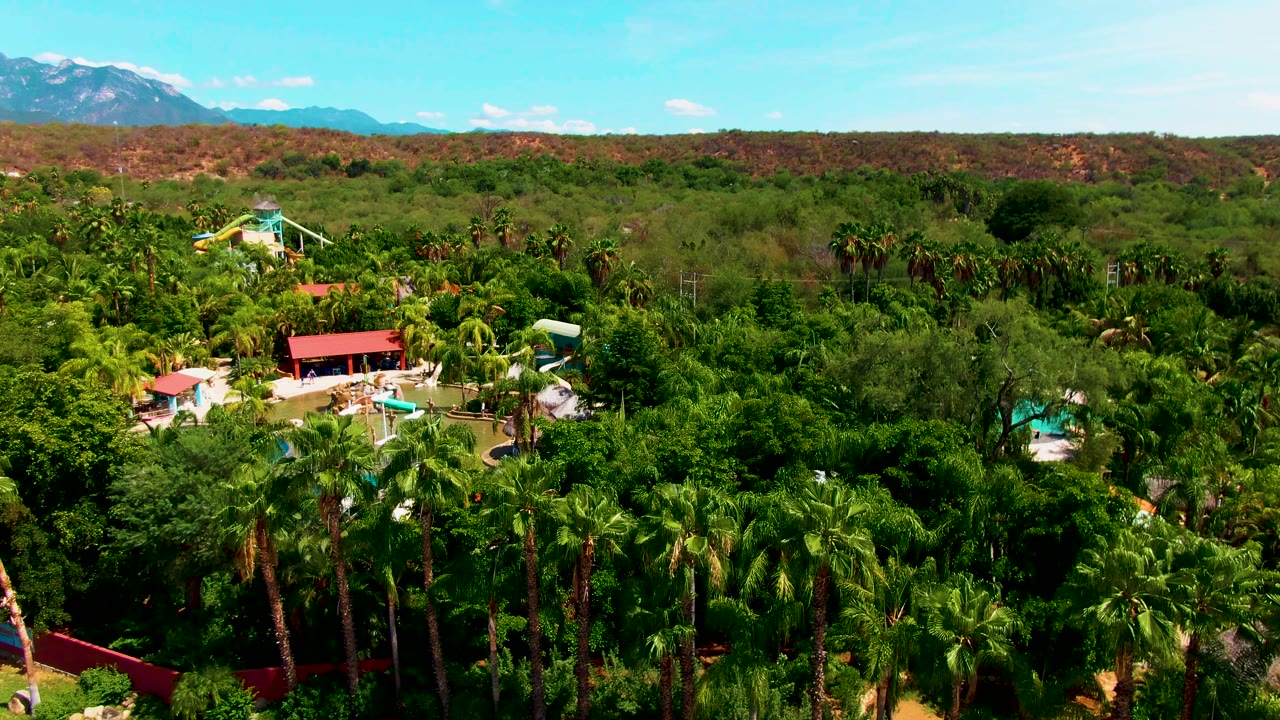 Aerial frame over a Caduano, Baja California Sur resort with palm canopy, small turquoise pools, red-roofed structures, pale water slides, reddish hills, distant mountains, and no visible people.