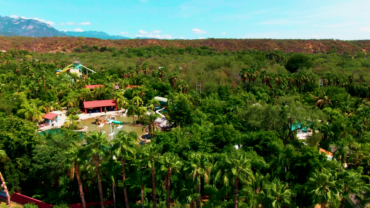 Aerial view in Caduano, Baja California Sur showing the pool complex slightly more centered among thick palm trees, with red roofs, white slide structures, desert ridge, distant mountains, and no visible people.