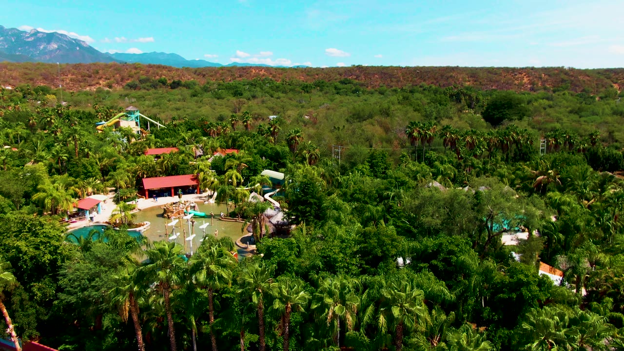 Wide aerial frame in Caduano, Baja California Sur with resort pools low in the image, palms filling the foreground and midground, pale slides, red roofs, a long reddish ridge, distant mountains, and no visible people.