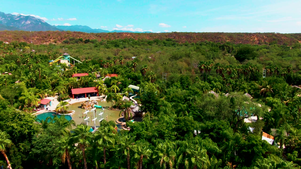 Palm-covered resort area with pool, slide, red roof, reddish hills, and mountains.