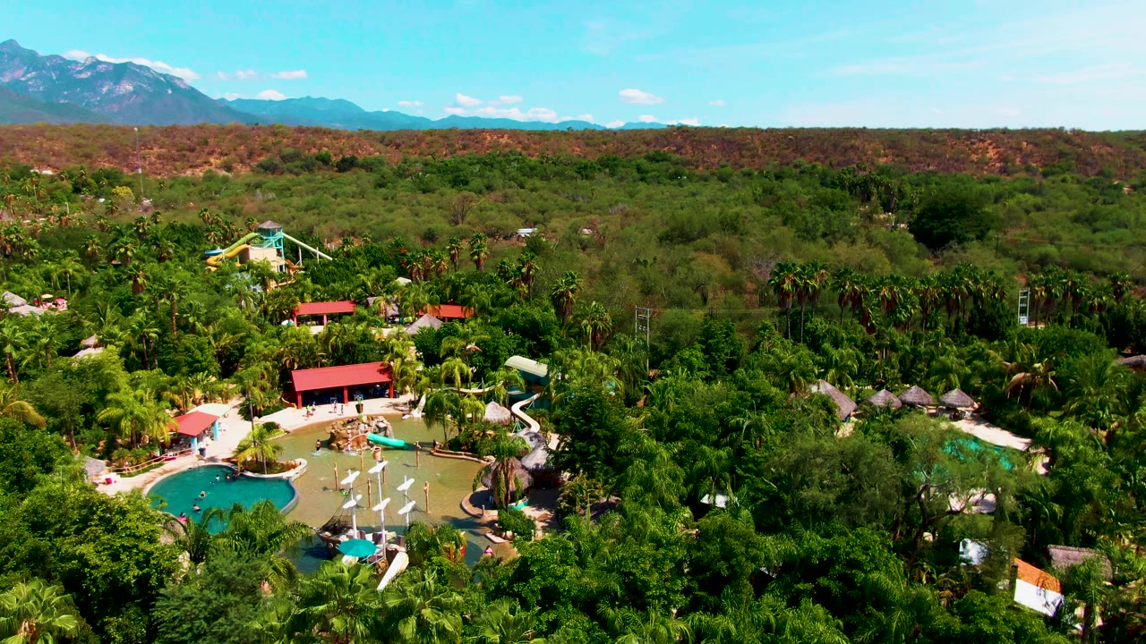 Aerial frame in Caduano, Baja California Sur showing turquoise resort pools, white umbrellas, red roofs, dense trees, a dry ridge, and distant mountains under blue sky.