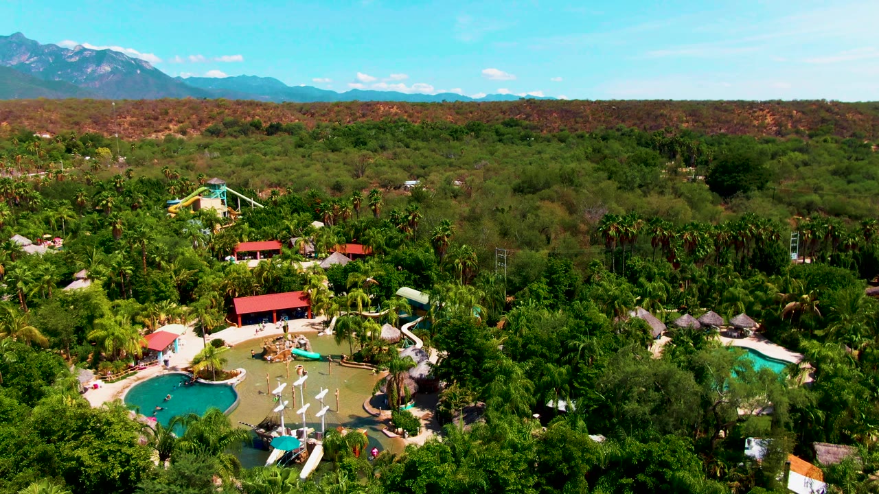 Caduano, Baja California Sur aerial frame showing resort pools and paths at the bottom edge, thick green vegetation in the middle distance, a reddish dry ridge, and blue mountains behind it.