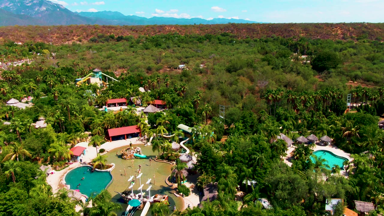 Second aerial frame in Caduano, Baja California Sur with the pool complex low in the view, thick palms and trees across the middle, reddish hills beyond, and no visible people.