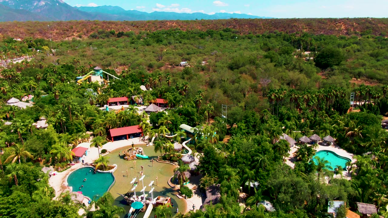 Aerial view over Caduano, Baja California Sur with bright blue pools, resort roofs partly surrounded by trees, a broad green canopy, reddish terrain, distant mountains, and no visible people.
