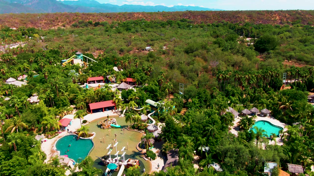 Later frame from the aerial clip in Caduano, Baja California Sur showing the pool area near the lower left, dense vegetation through the center, dry reddish hills, mountains, and no visible people.