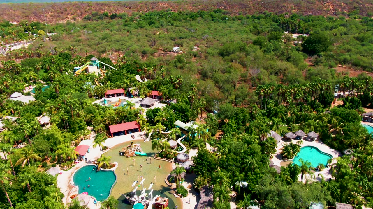 Aerial view over a Caduano resort area with turquoise pools, palm trees, red-roof structures, dry hills in the distance, and no visible people.
