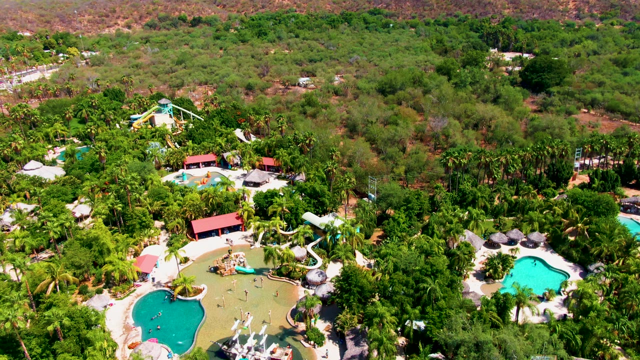 High aerial frame in Caduano showing multiple pool shapes, red-roof resort buildings, thick palms, hillside vegetation, and no visible people.