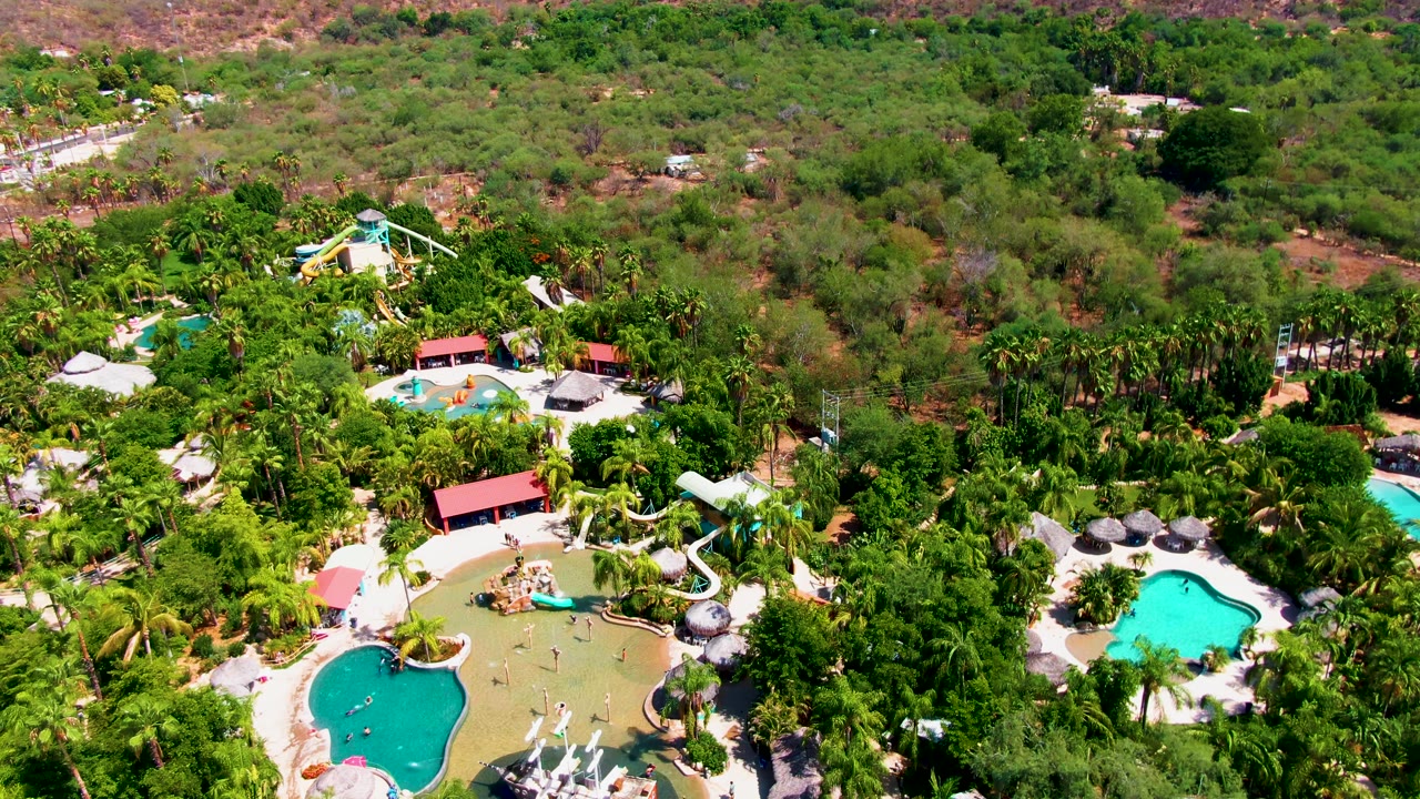 Drone-height view of the Caduano resort grounds with bright pool water, clustered palms, light-colored paths, red roofs, and dry green-brown hills beyond, with no visible people.
