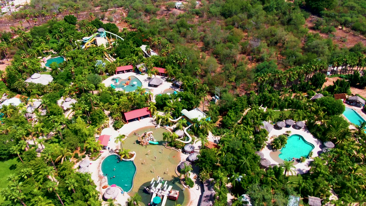 Aerial view in Caduano, Baja California Sur showing multiple turquoise pools, a white water slide, resort roofs, umbrellas, and dense greenery at the opening moment.
