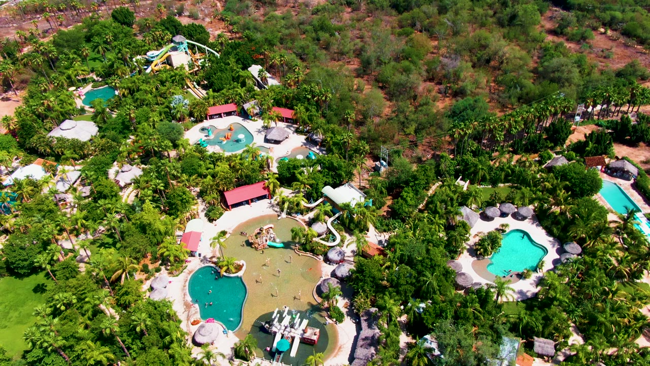 Aerial frame over Caduano, Baja California Sur showing the white slide feeding toward a pool, clustered umbrellas, bright turquoise water, and planted greenery filling most of the view.