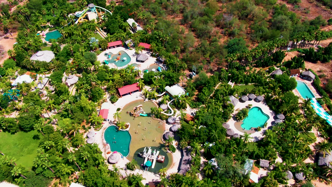 Overhead view of resort pools, white and blue water slides, palm-like greenery, and dry terrain in Caduano, Baja California Sur, with no visible people.