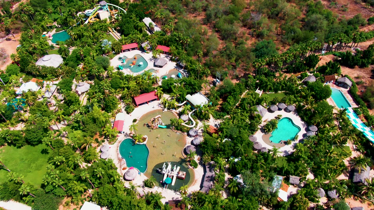 Aerial frame over Caduano resort grounds with the main pool cluster lower left, curved white slides near center, blue slide lanes at right, and no visible people.