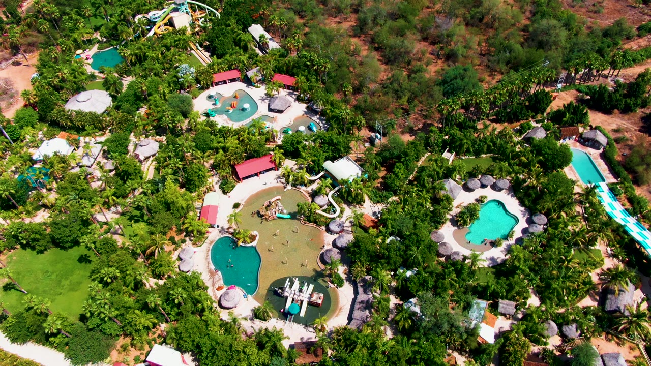 Top-down resort water park view in Caduano with green planting around several pools, pale paths, red roofs, blue slide lanes on the right, and no visible people.