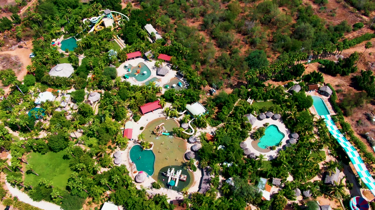 Overhead view of a Caduano, Baja California Sur water park with turquoise pools, curved slides, red-roof structures, dense trees, dry terrain beyond the grounds, and no visible people.