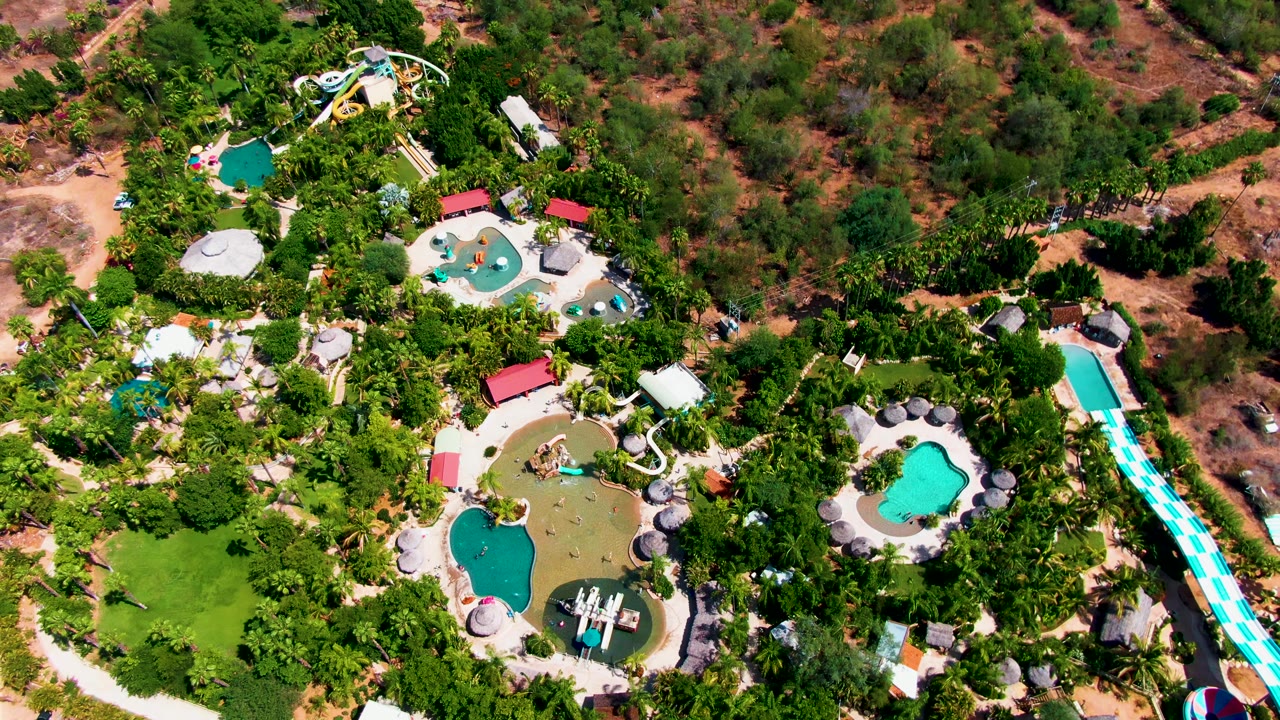 Aerial frame of the Caduano water park showing the long blue-and-white slide more prominently at right, clustered pools in the center, surrounding vegetation, and no visible people.