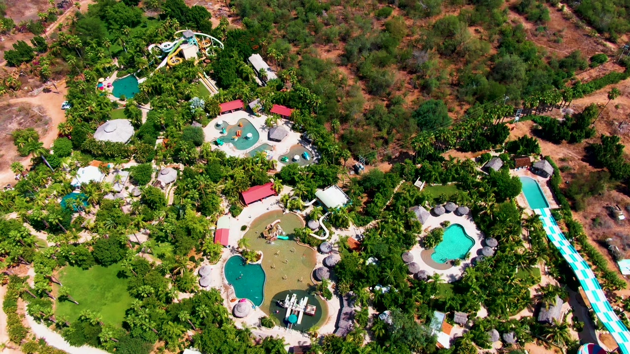 High overhead view in Caduano, Baja California Sur with multiple pools, pale paths, a curved white slide, dense tropical planting, dry scrubland at the top edge, and no visible people.