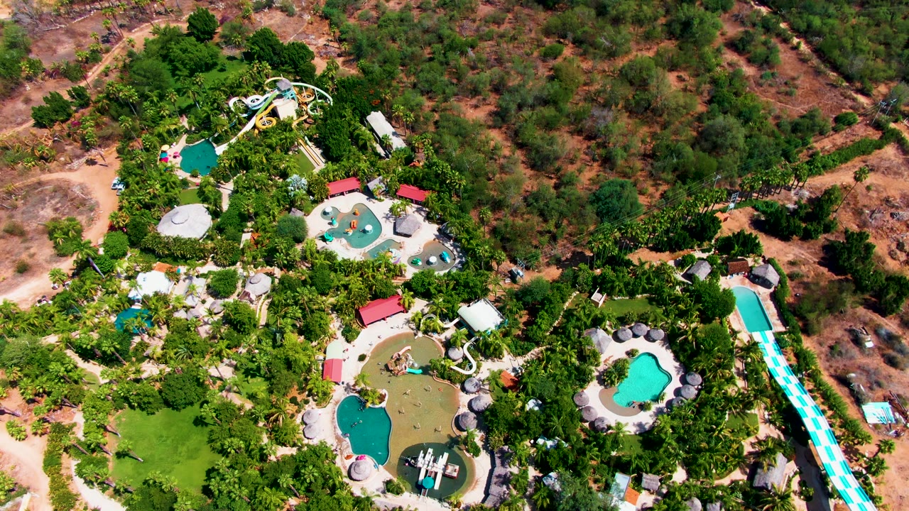 High aerial frame over a Caduano, Baja California Sur water park, with turquoise pools, winding slides, dense palms, dirt access roads, and dry brushland around the property.