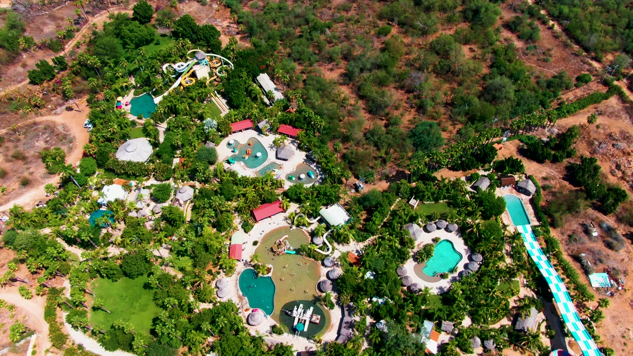 Overhead Caduano water park frame with several pools clustered among palms, a long blue-and-white striped slide along the right side, and brown open ground above the park.
