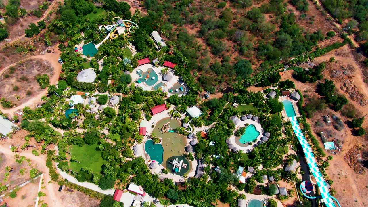 High overhead view of a Caduano, Baja California Sur water park with turquoise pools, a long striped slide at right, dense trees, dry terrain, and no visible people.