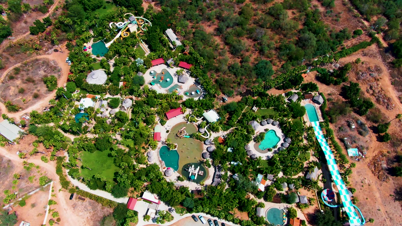 High drone view over Caduano, Baja California Sur with multiple pools clustered among trees, a blue-and-white slide near the right edge, dry scrubland around the park, and no visible people.