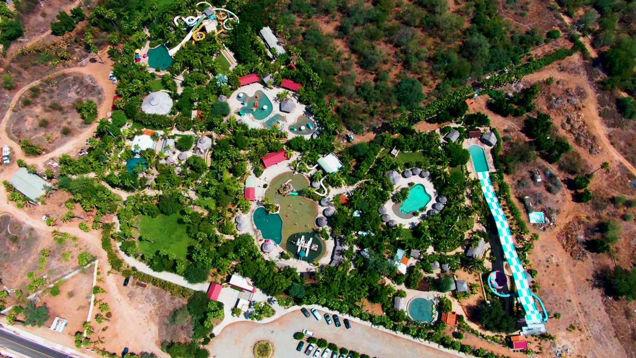Overhead aerial frame of a water park in Caduano, Baja California Sur, Mexico, showing turquoise pools, a long straight slide, parked cars, a paved road, trees, and dry open land with no visible people.