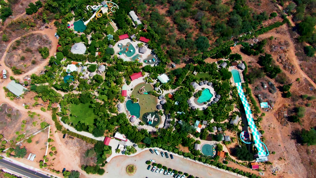 Aerial frame over Caduano water park with multiple blue-green pools, branching walkways, a tall slide on the right edge, parked vehicles below, and brown hillside terrain around the property with no visible people.