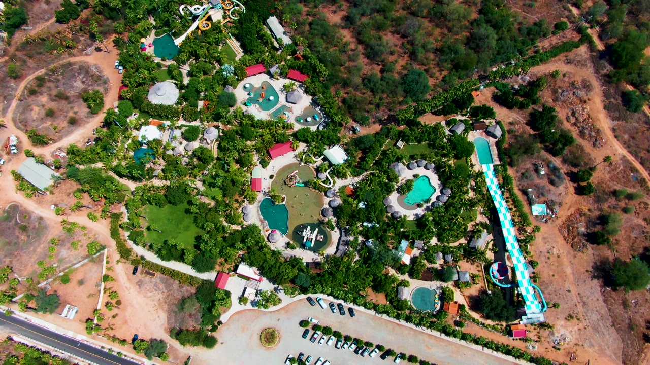 Top-down view of the Caduano, Baja California Sur water park, with the long slide running near the right side, clustered pools and trees in the center, cars in the lower parking area, and no visible people.