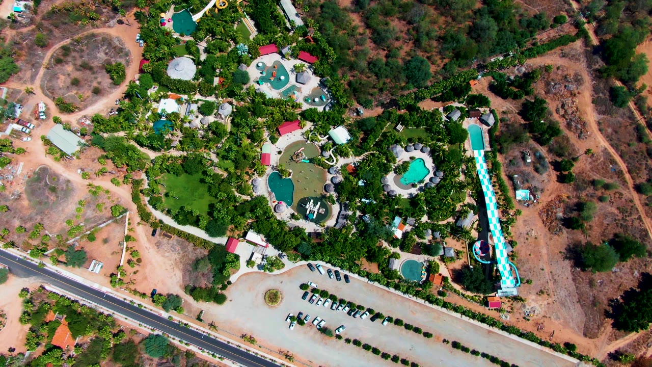 Overhead view in Caduano, Baja California Sur showing resort pools, a long water slide, parking rows, a diagonal road, surrounding greenery, dry hillside, and no visible people.