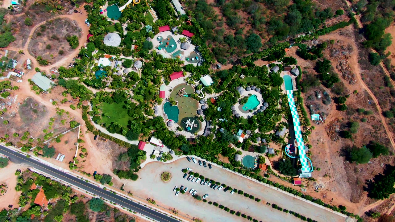 Aerial frame over Caduano, Baja California Sur with multiple turquoise pools, dense trees, parked cars, a roadside entrance area, dry open ground, and no visible people.
