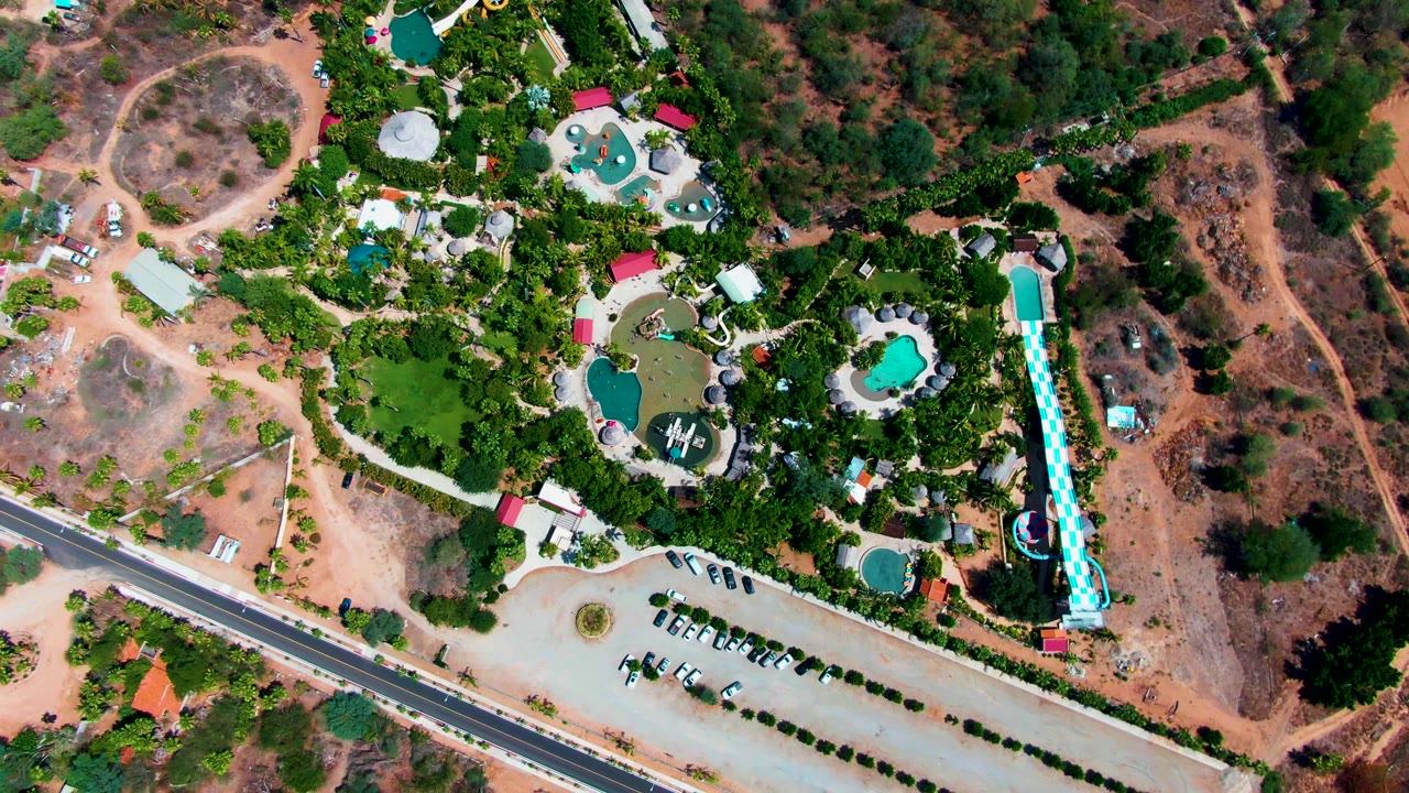 Straight-down view of a Caduano resort water park in Baja California Sur, with a tall blue-and-white slide, rounded pools, red roofs, parking rows, scrubland, and no visible people.