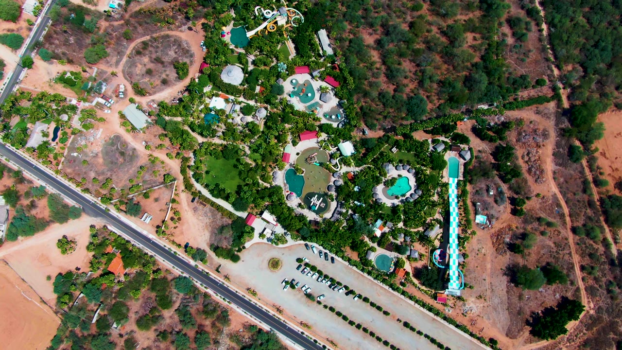 Aerial frame in Caduano, Baja California Sur, with the paved road cutting past a green resort complex, turquoise pools, a tall water slide, parking rows, dry ground, and no visible people.