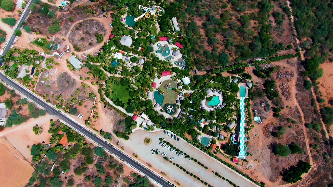 Drone frame in Caduano, Baja California Sur, showing resort pools and slide structures beside a paved road, with dry lots and darker tree cover around the developed area and no visible people.