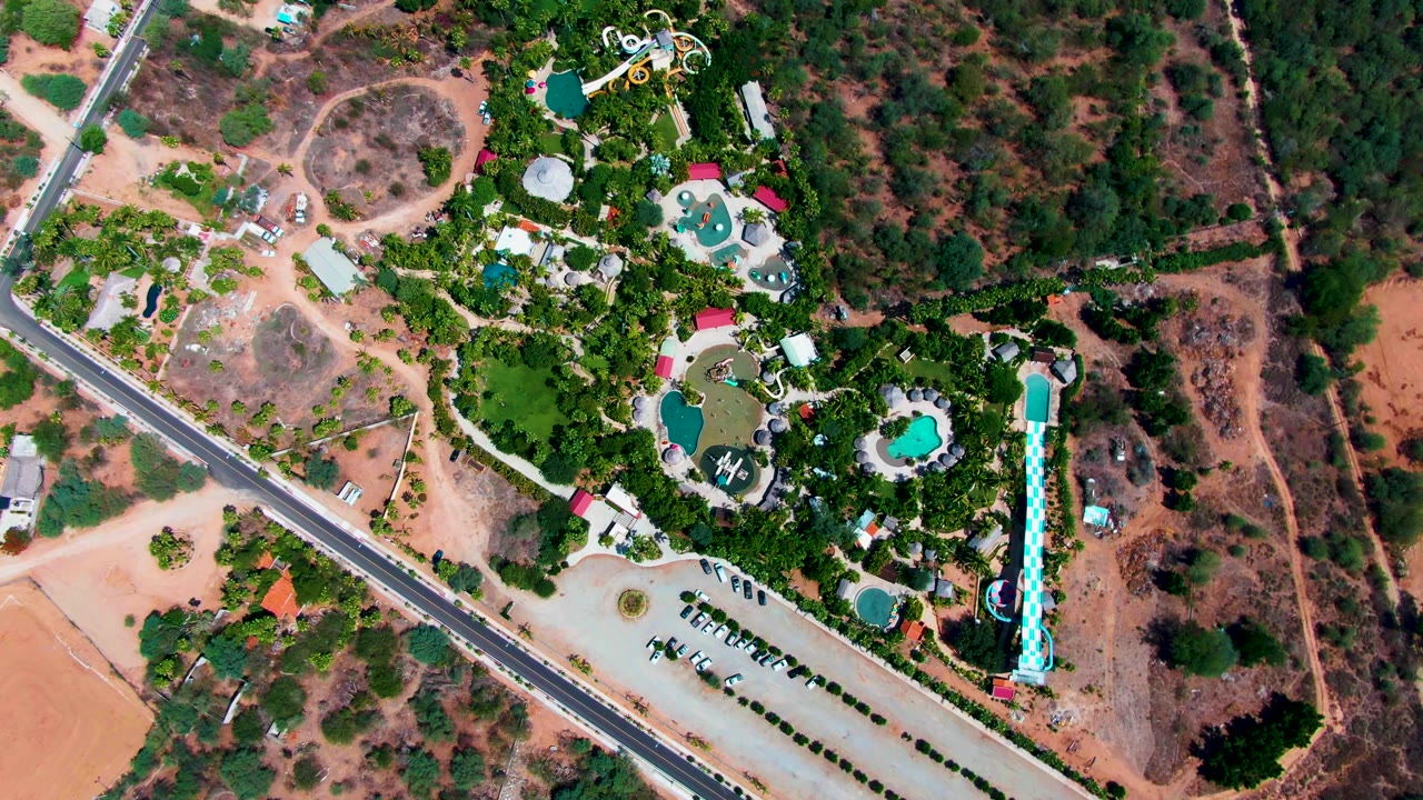 Overhead frame in Caduano, Baja California Sur, with the road along the left side, a parking area below the resort, clustered pools and slides in greenery, dry hillsides, and no visible people.