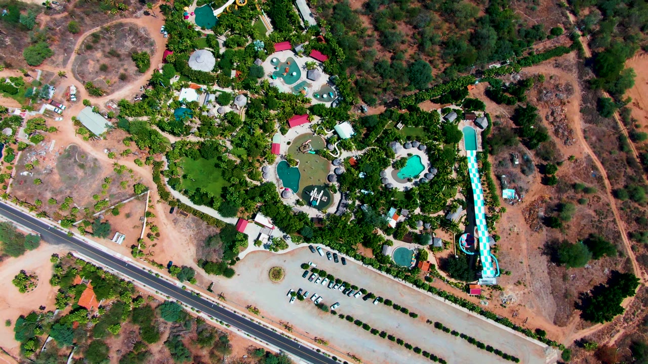 Top-down aerial view of a resort in Caduano, Baja California Sur, with turquoise pools, a long blue slide, a paved road, parked vehicles, green trees, dry brown ground, and no visible people.