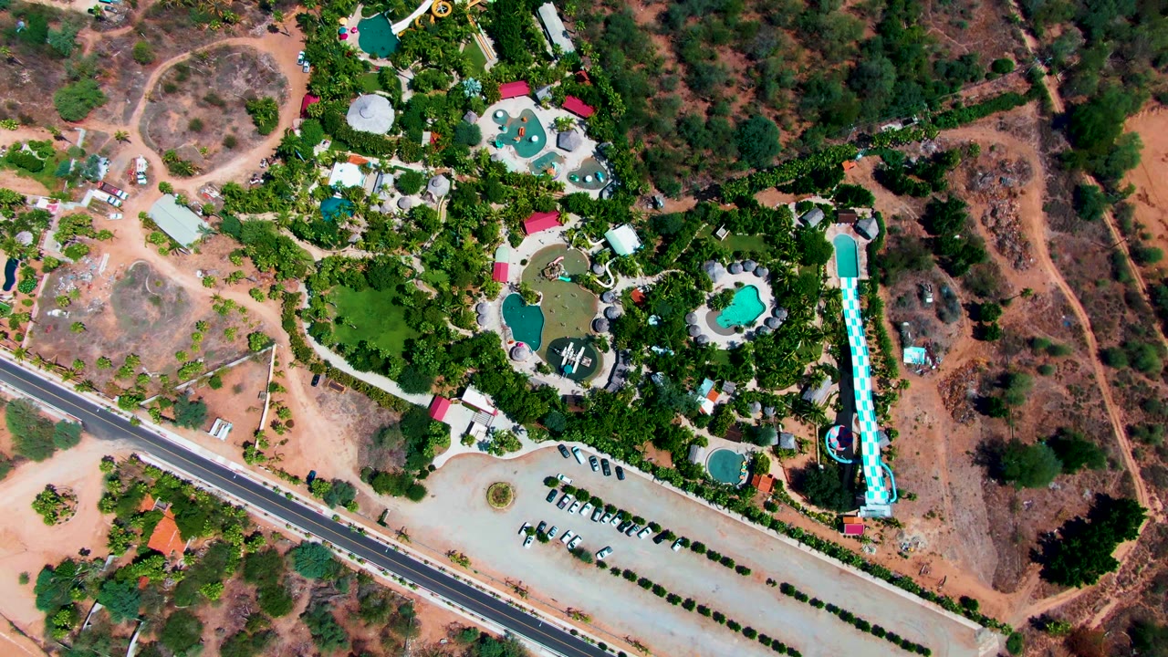 Overhead frame of the Caduano resort grounds showing the pool cluster near dense greenery, the tall blue slide on the right, a pale parking area with vehicles, a paved road, and no visible people.