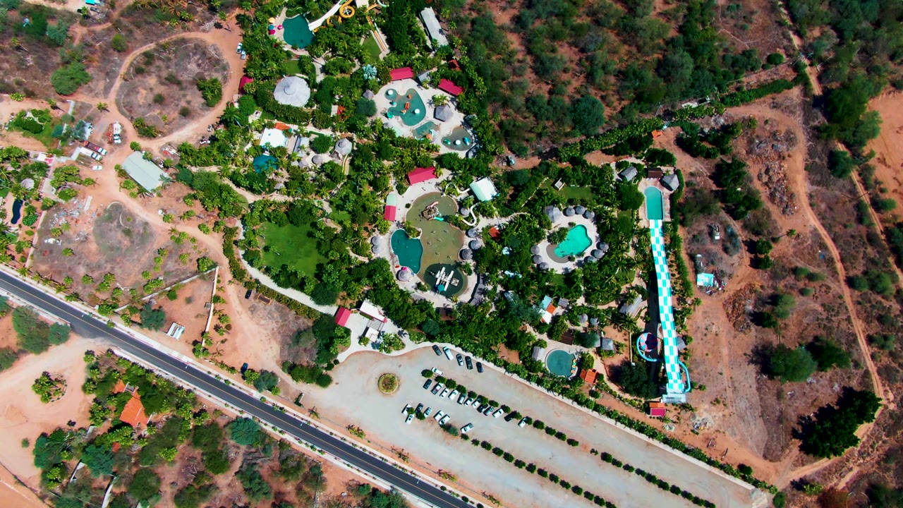 Aerial overhead view in Caduano, Baja California Sur, with bright blue pools and slide set among trees, a row of parked vehicles below, a paved road to the left, dry open land to the right, and no visible people.