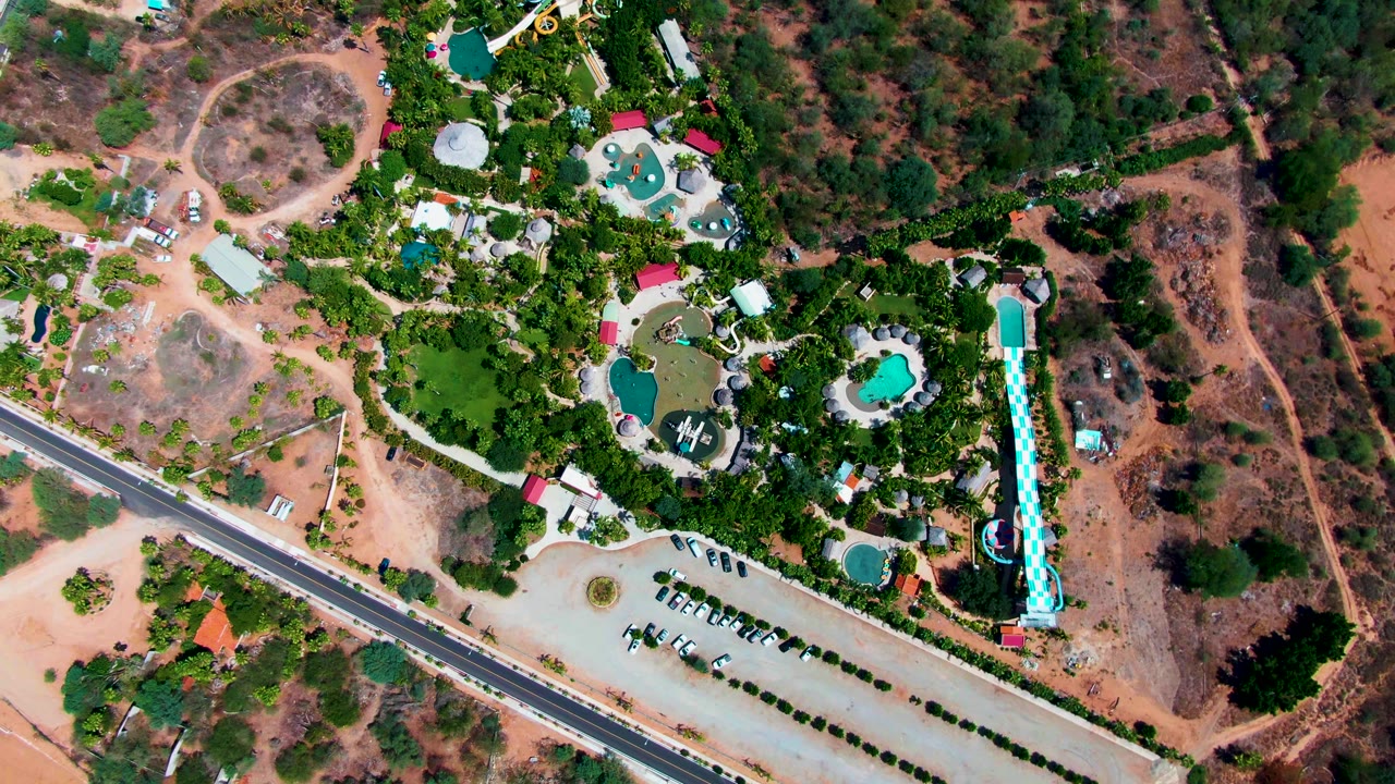 Top-down drone view of the Caduano resort area with multiple pools, the blue slide along the right side, planted green areas, parked vehicles in a pale lot, the road at lower left, and no visible people.
