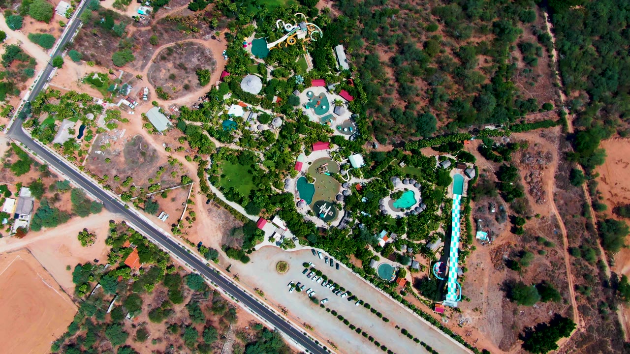 High overhead view of a Caduano, Baja California Sur resort with turquoise pools, a long slide, diagonal road, parking area, trees, dry reddish land, and no visible people.