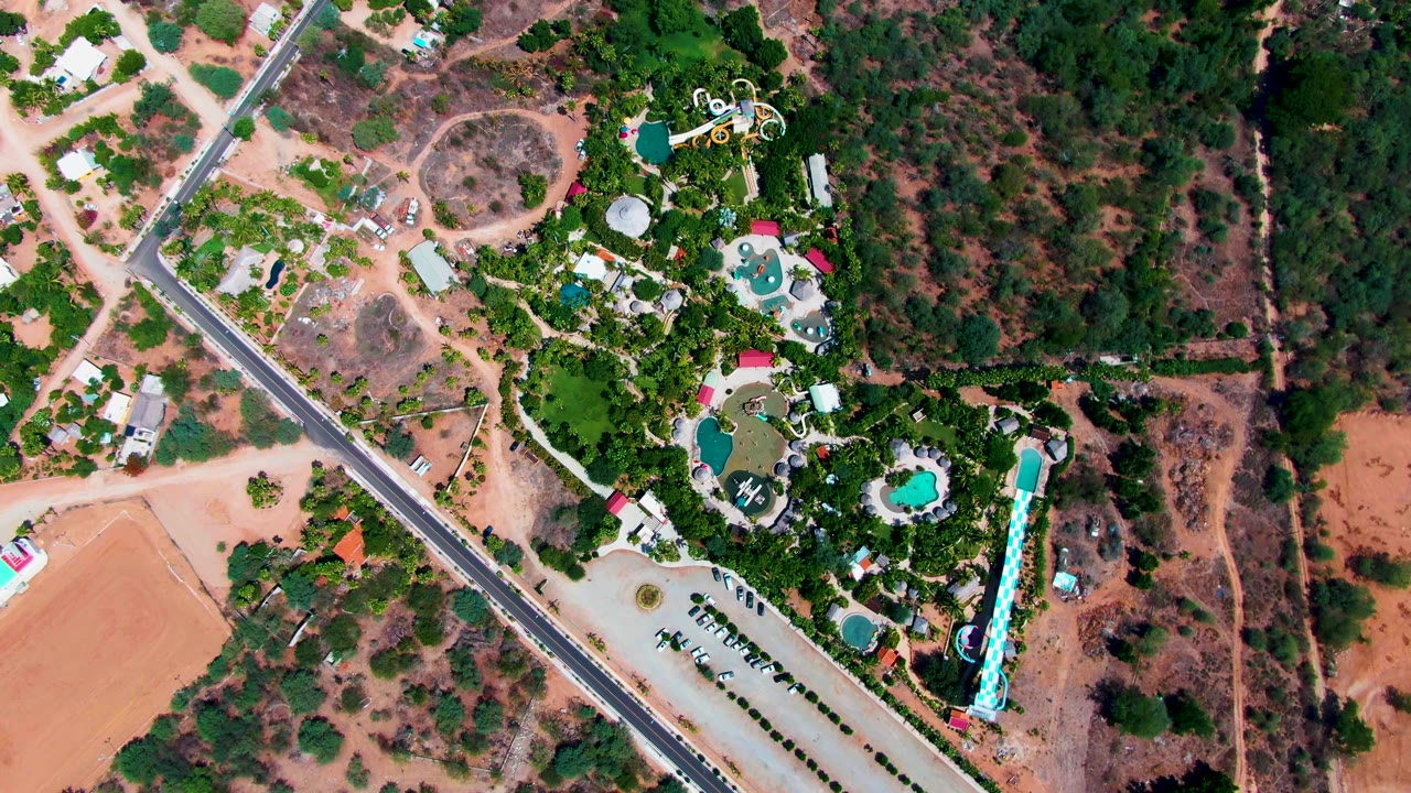 High aerial view in Caduano, Baja California Sur showing a resort with blue pools, green trees, parking areas, a diagonal road, dry open land, and no visible people.