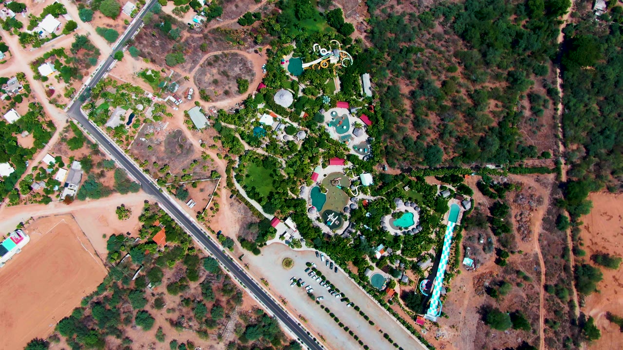 Overhead Caduano resort frame showing multiple blue pool shapes among dense greenery, a road cutting diagonally through the left side, dry brushland to the right, and no visible people.