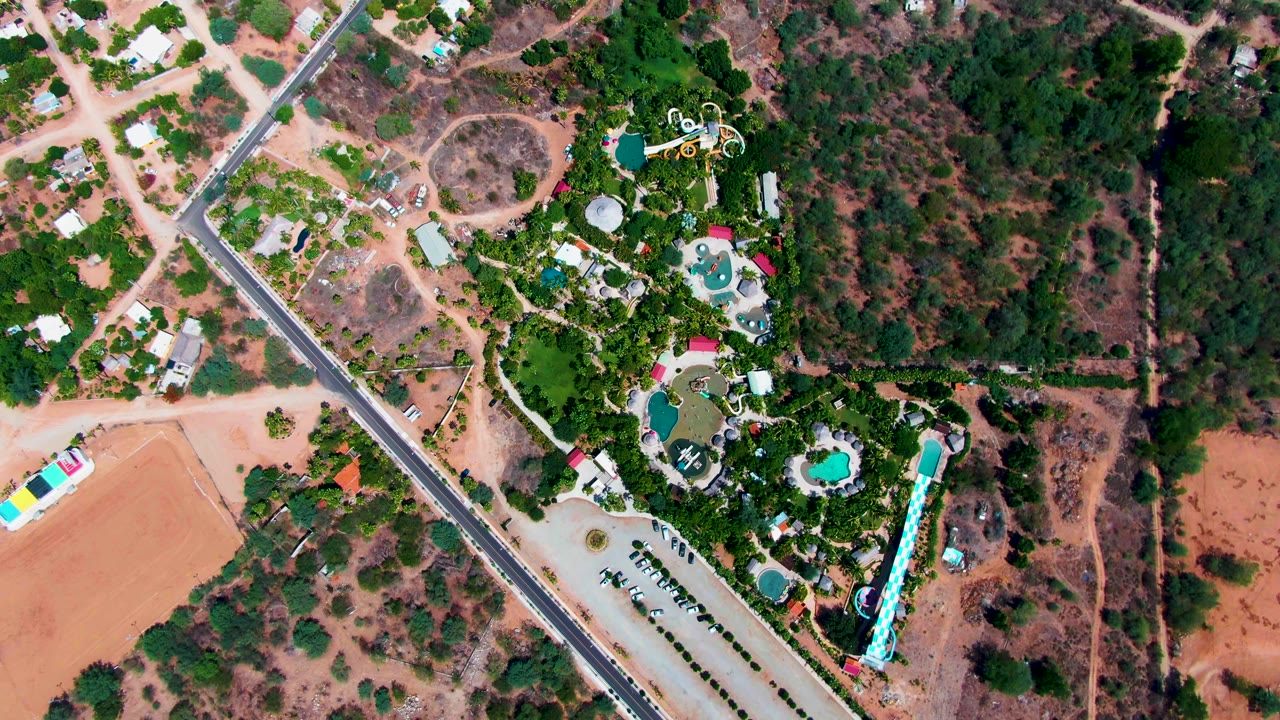 Overhead view of Caduano, Baja California Sur with a diagonal road, houses, blue pools, dry lots, green trees, and a small white vehicle near the road, no visible people.