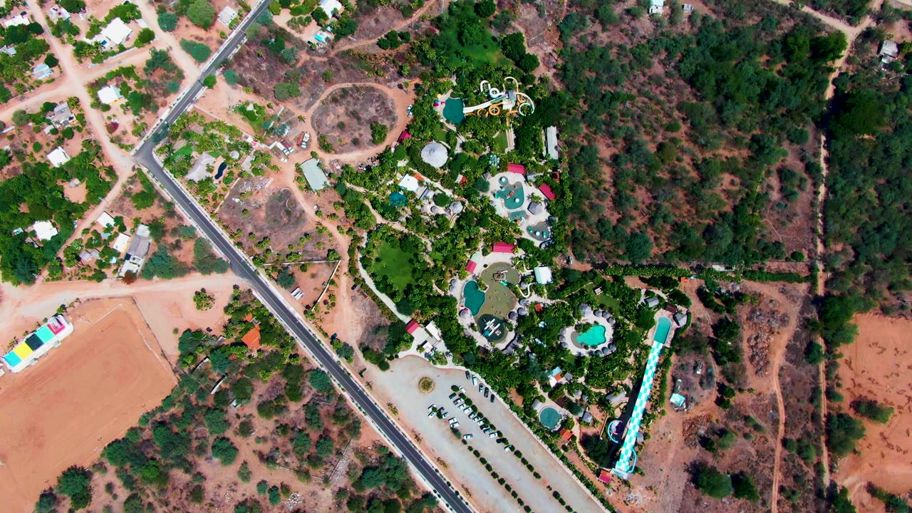 Aerial view of diagonal road beside houses, pools, dirt fields, and dense trees