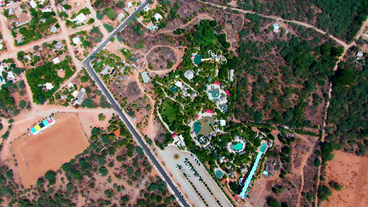 Overhead drone frame in Caduano, Baja California Sur, showing a diagonal paved road, residential buildings, green landscaping, pools, and reddish dry ground with no visible people.