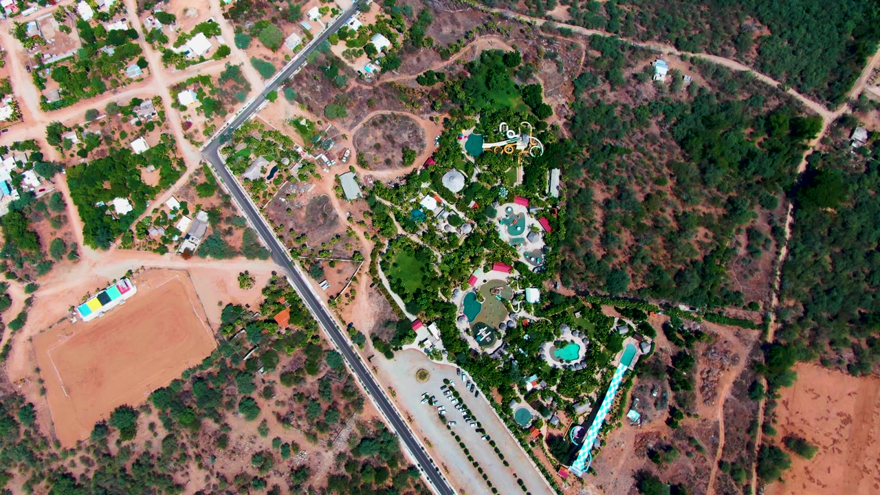 Second overhead frame in Caduano, Baja California Sur, with the straight road crossing the left side, clustered homes and trees above it, and pool areas set into dense greenery, no visible people.