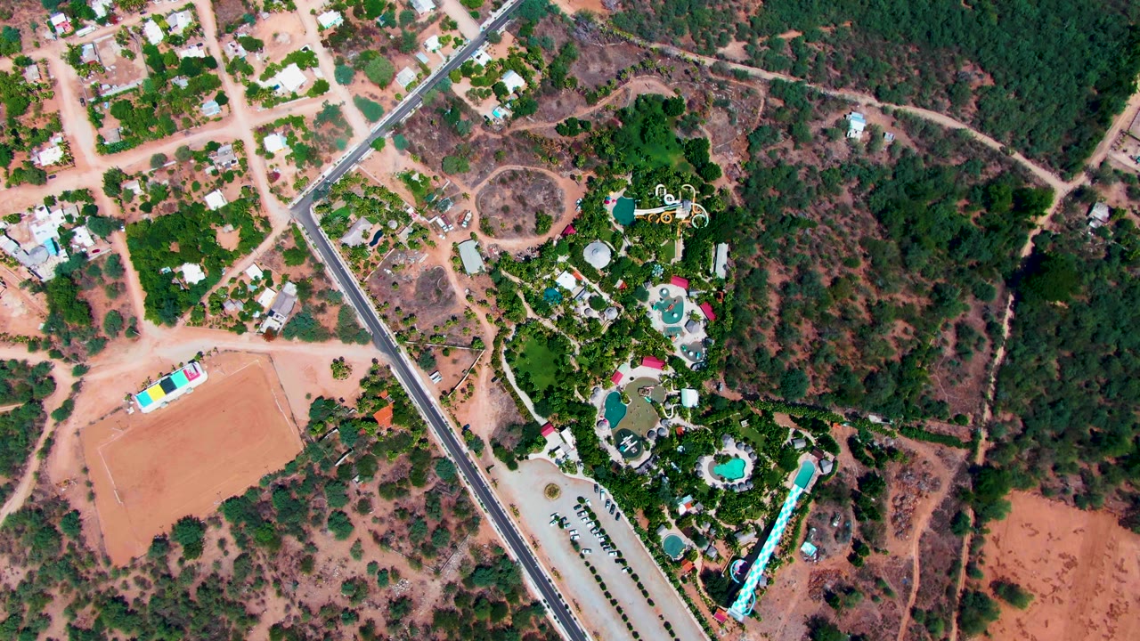 High overhead frame of Caduano, Baja California Sur showing a tan dirt field, paved road, scattered residential structures, dense trees, and resort-style pools with no visible people.