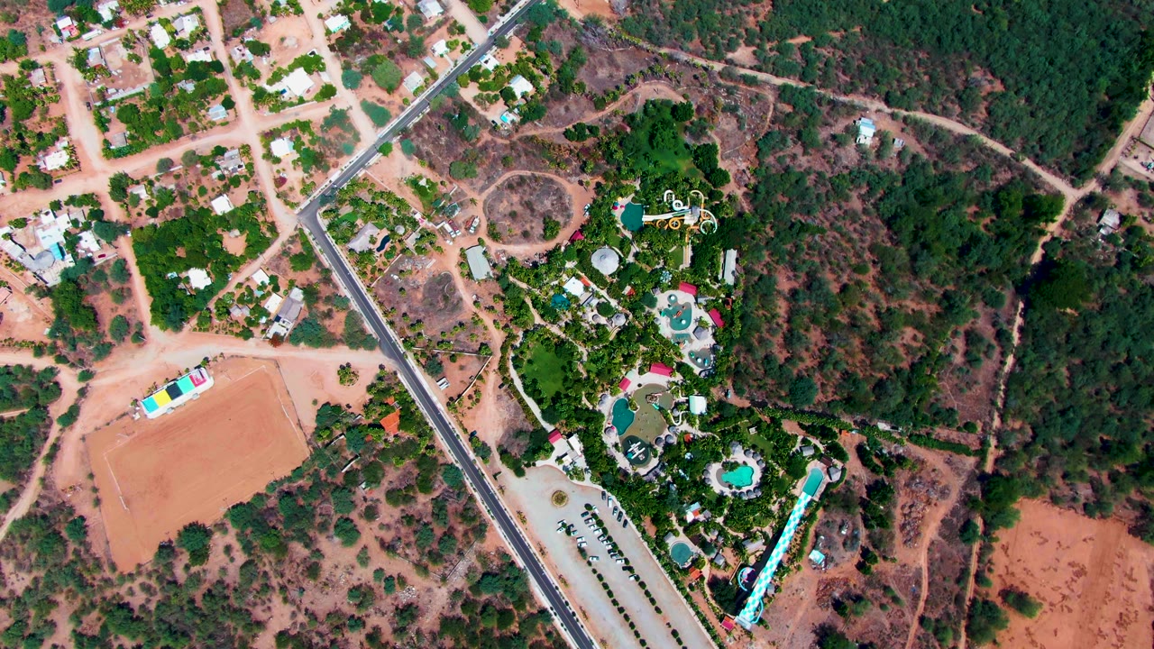 Second frame of the Caduano aerial keeps the dirt field on the left, the paved road near center, and the landscaped green area with pools on the right, with no visible people.
