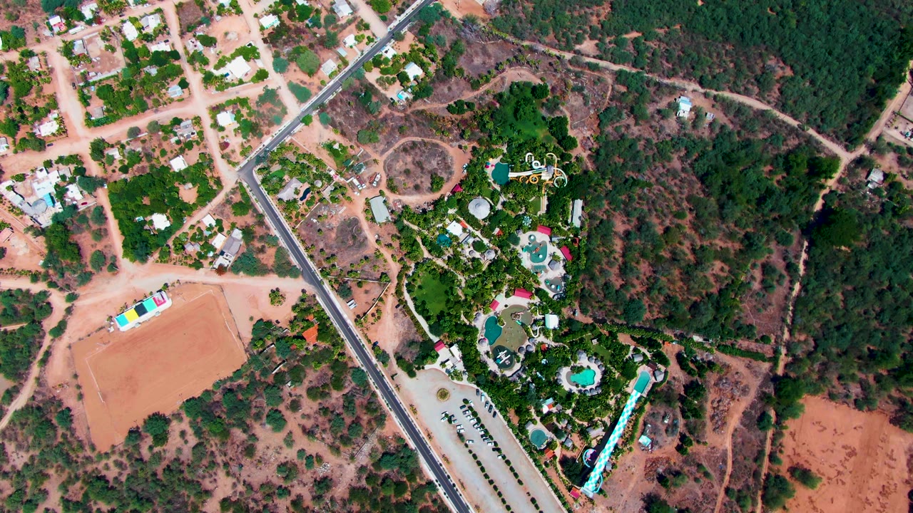 Mid-clip aerial frame over Caduano showing pale dirt roads and lots at top left, a main paved road, dry scrub, and a compact green area with pools, with no visible people.
