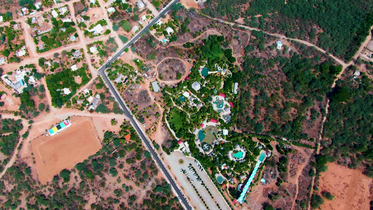 Overhead view of a dirt field, road, residences, trees, and pools.
